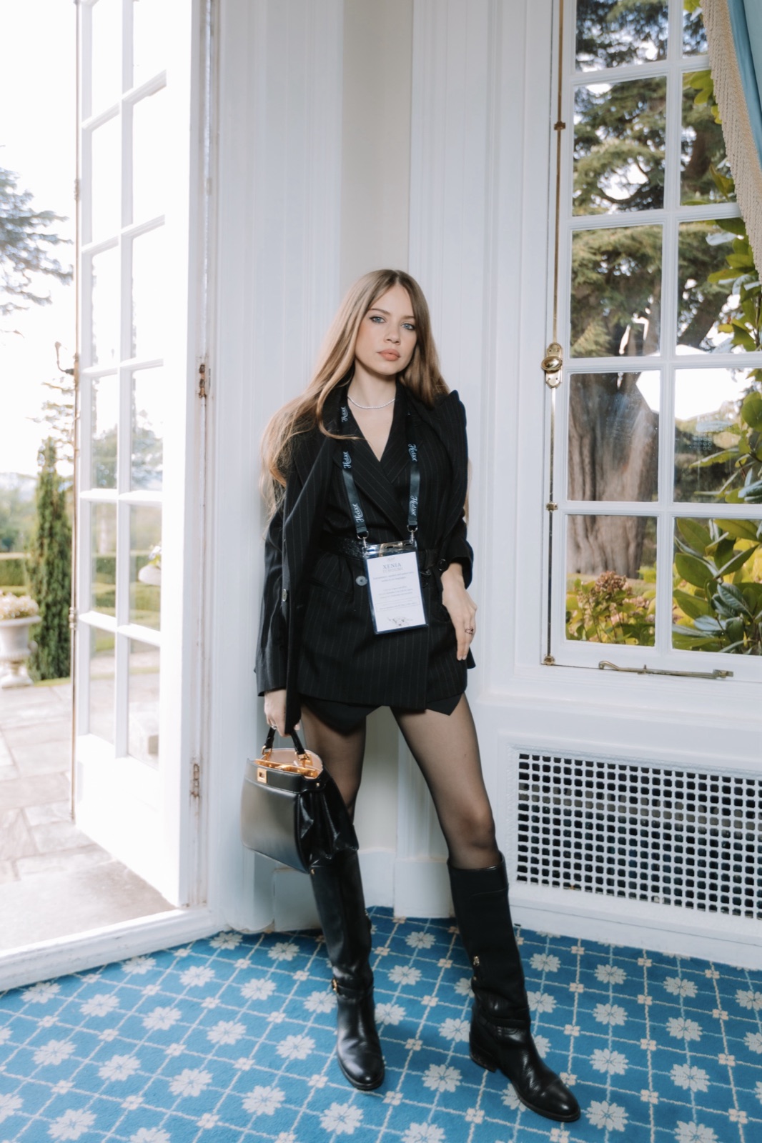 Xenia Tchoumi at a window of Hedsor House wearing her Hedsor Hundred name card, in a Windsor pinstripe suit, Ralph Lauren knee boots and a black Fendi bag, The Hedsor Hundred 2026