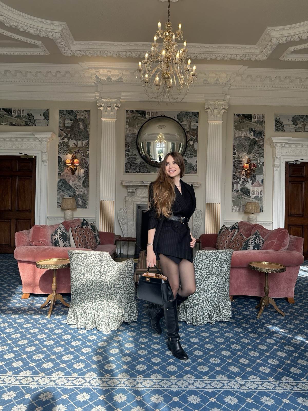 Xenia Tchoumi in the drawing room of Hedsor House at The Hedsor Hundred 2026, in a Windsor pinstripe suit, Ralph Lauren knee boots and a Fendi bag beneath a crystal chandelier