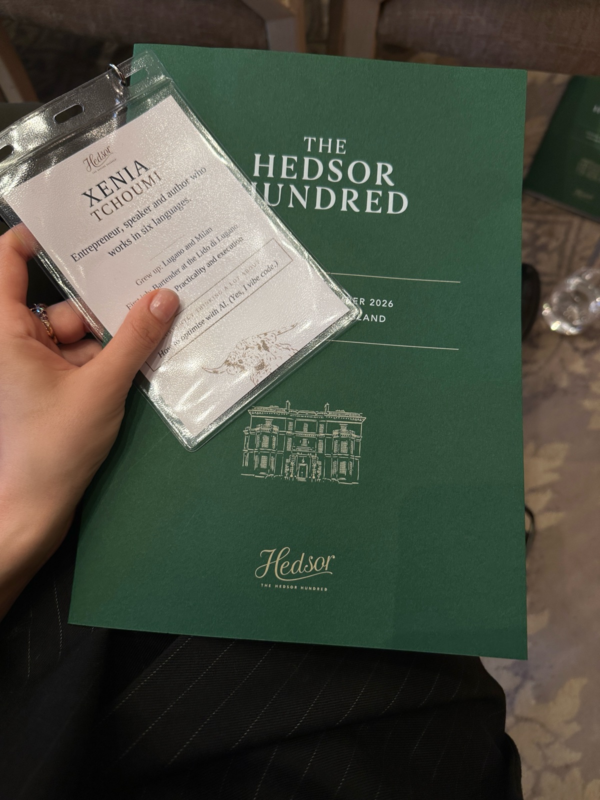 Xenia Tchoumi's name card for The Hedsor Hundred, describing her as an entrepreneur, speaker and author who works in six languages, on the engraved green programme